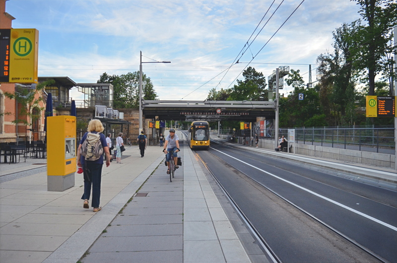 7 - An urban street scene featuring a public transit tram stop with a yellow tram approaching on tracks alongside a paved pedestrian sidewalk and a dedicated bicycle lane